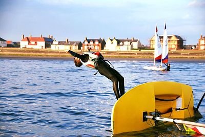 Young sailor in a wetsuit launching off the bow of a dinghy on a sunny day at Prestwick, with another dinghy and the coastline in the background